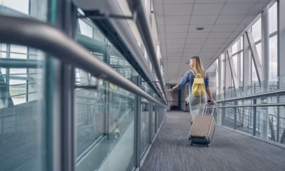 Mulher de costas caminhando por um corredor envidraçado de aeroporto, carregando uma mochila amarela e puxando uma mala de rodinhas durante uma conexão ou escala de voo.