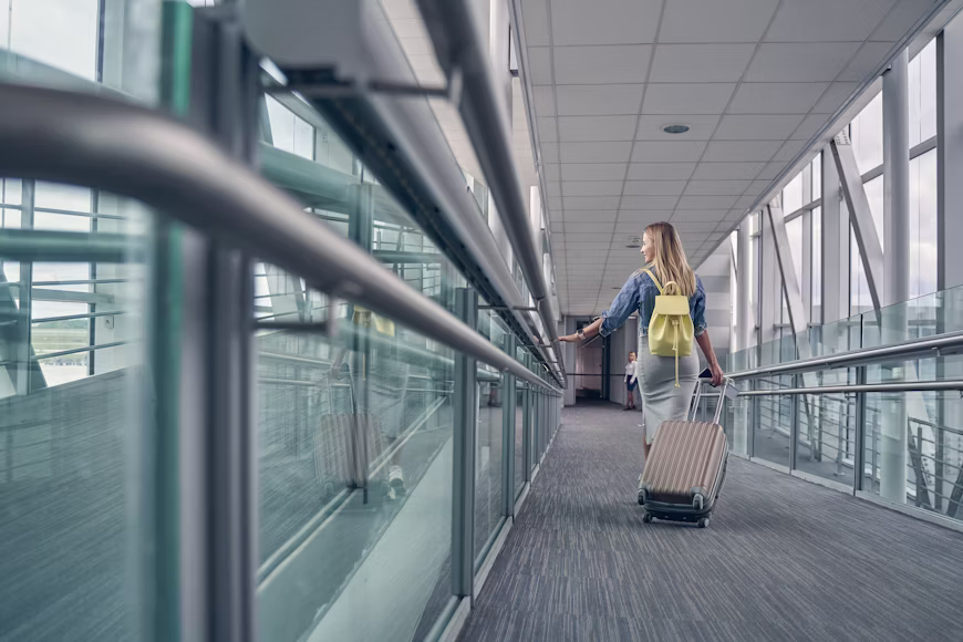 Mulher de costas caminhando por um corredor envidraçado de aeroporto, carregando uma mochila amarela e puxando uma mala de rodinhas durante uma conexão ou escala de voo.