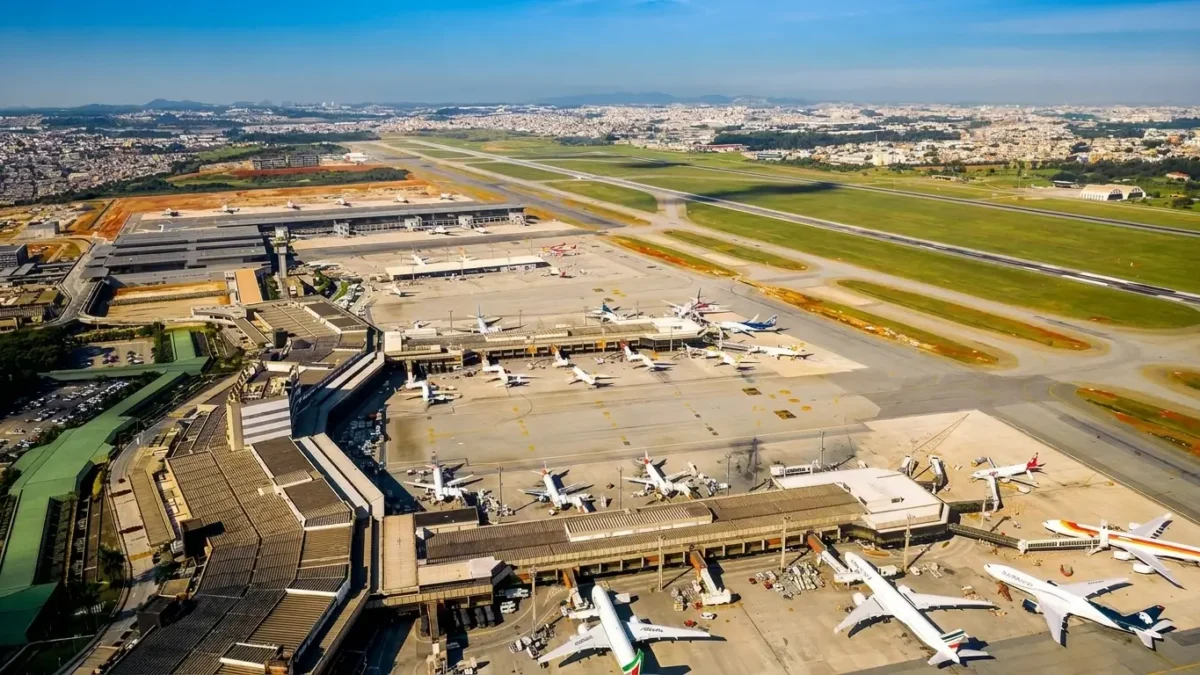Vista aérea do Aeroporto Internacional de Guarulhos com terminais, pistas e aviões estacionados em um dia ensolarado.