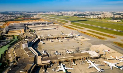 Vista aérea do Aeroporto Internacional de Guarulhos com terminais, pistas e aviões estacionados em um dia ensolarado.