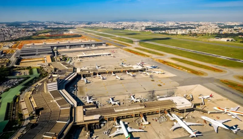 Vista aérea do Aeroporto Internacional de Guarulhos com terminais, pistas e aviões estacionados em um dia ensolarado.