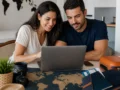 Como comparar preços de pacotes de viagem sem cair em falsas ofertas - destacada: Casal comparando preços de pacotes de viagem em notebook com mapas e documentos de viagem